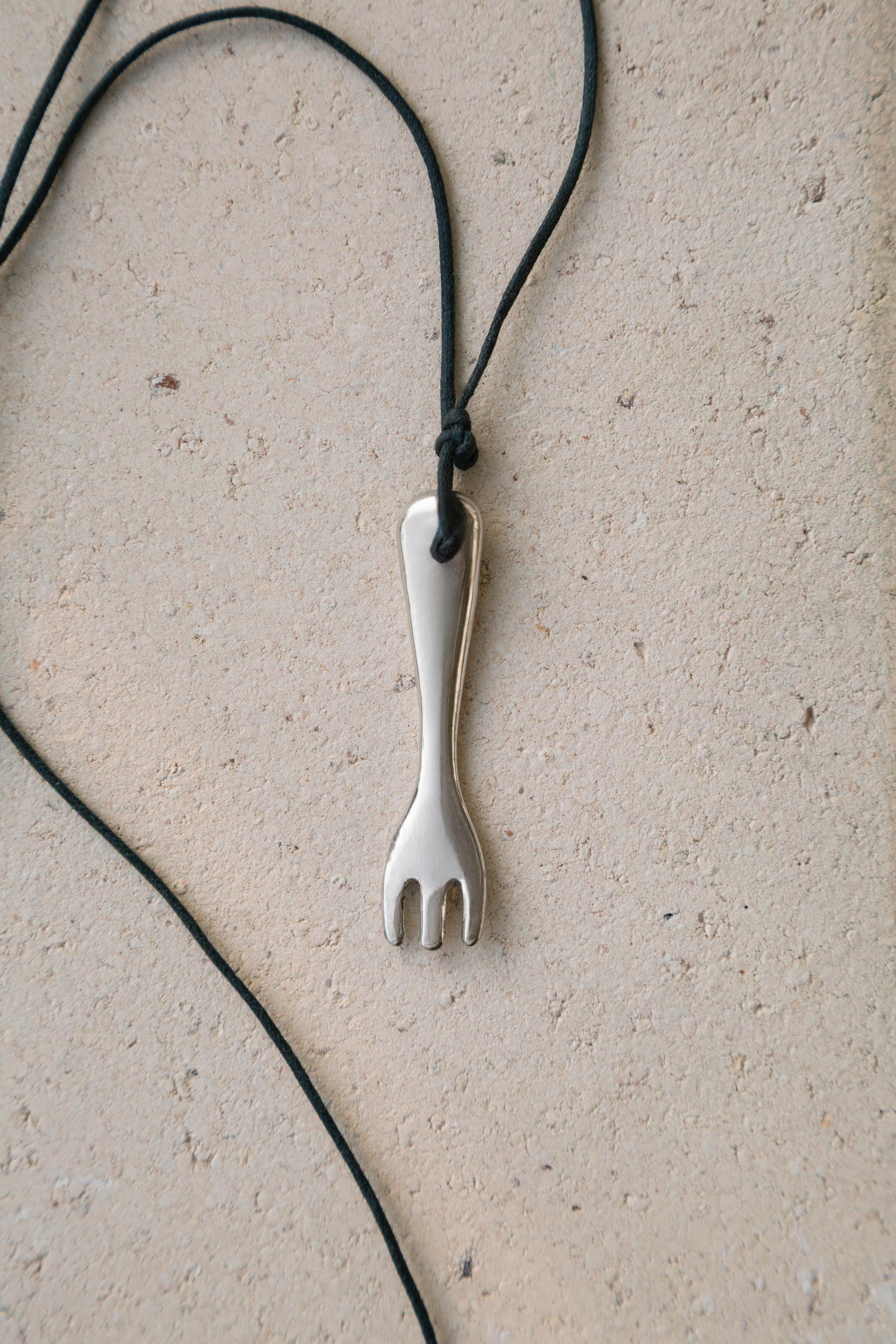 A silver metal tasting fork pendant necklace on a black cord, laid flat on a textured beige surface.