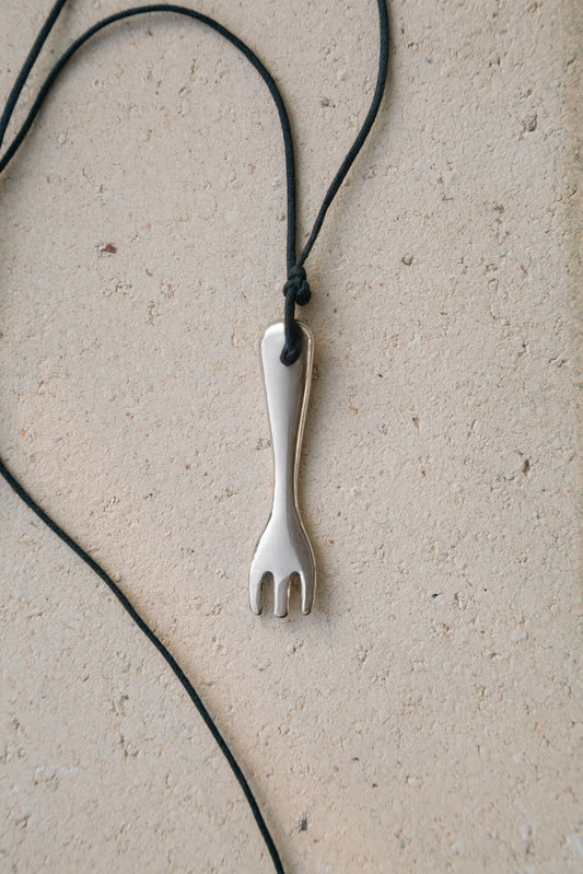 A silver metal tasting fork pendant necklace on a black cord, laid flat on a textured beige surface.